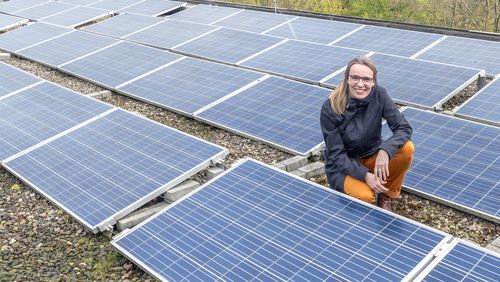 Eine Frau hockt auf einem Flachdach, das mit Solarmodulen bedeckt ist und lächelt in die Kamera.
