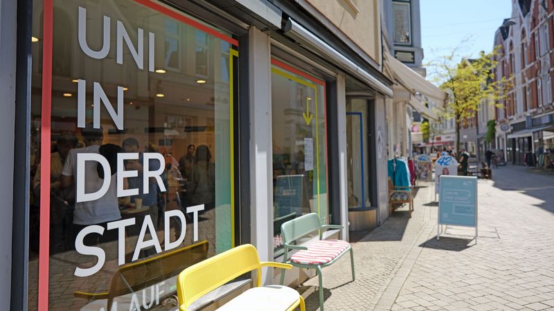 Das Bild zeigt den Store von außen. Auf dem Schaufenster sowie auf einem Aufsteller auf der Straße steht der Schriftzug "Uni in der Stadt". Vor dem Geschäft stehen bunte Bänke mit Kissen. Der Himmel im Hintergrund ist blau, die Sonne scheint.