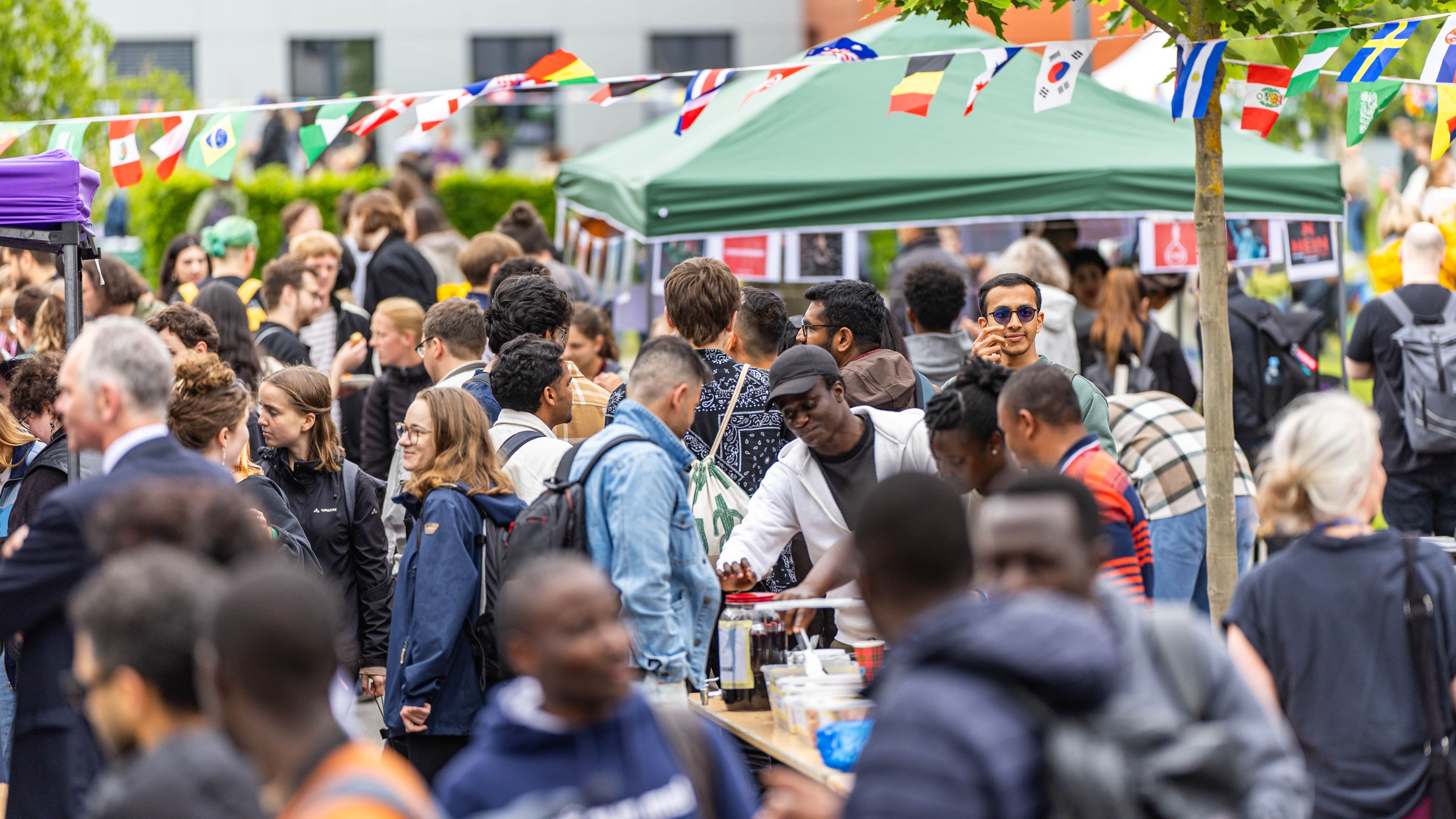 The picture shows visitors to the International Summer Fest. They are walking around, standing at the stands, chatting or eating and drinking. A chain with different national flags hangs above the square.