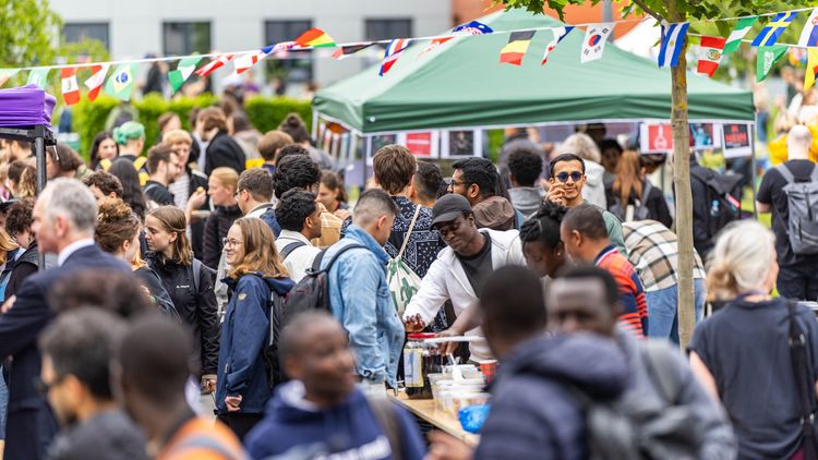 The picture shows visitors to the International Summer Fest. They are walking around, standing at the stands, chatting or eating and drinking. A chain with different national flags hangs above the square.