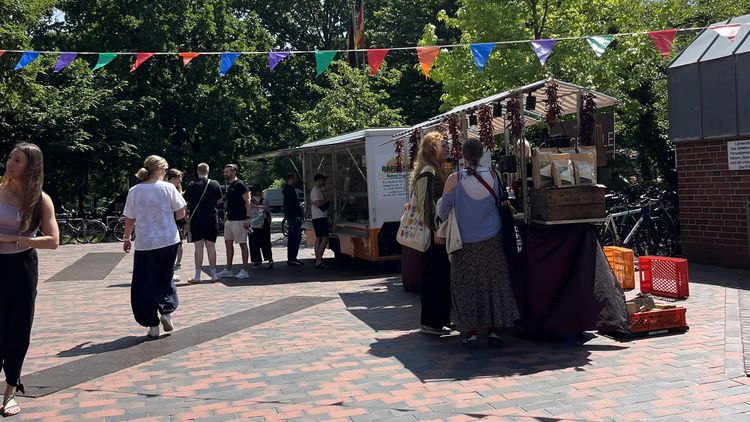 Das Bild zeigt ein paar Verkaufsstände während des Campusmarktes auf dem Mensavorplatz. Menschen stehen um sie herum und schauen sich die Produkte an. Eine Leine mit bunten Fähnchen hängt über dem Platz. Der Himmel ist blau, die Sonne strahlt.