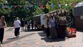 The picture shows some stalls at the Campus Market on the Mensa forecourt. People are standing around them and look at the products. A line of colourful flags hangs over the square. The sky is blue, the sun is shining.