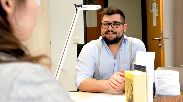 Max Bauer sitzt mit einer Studentin an einem Tisch, im Vordergrund sind einige Bücher zu sehen.