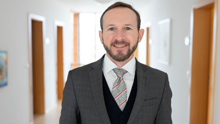 The picture shows Johannes Lorenz. He stands in a corridor next to a banister and smiles at the camera. The corridor and office doors can be seen in the background.