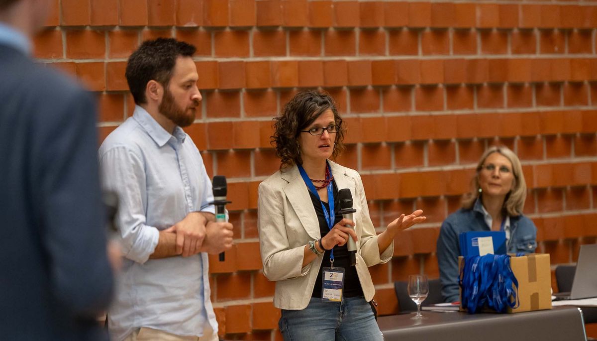 Symposium Zwei Personen stehen vor einer roten Backsteinwand und sprechen in Mikrofone. Eine Frau in einem beigen Blazer gestikuliert lebhaft, während ein Mann neben ihr mit einem Mikrofon in der Hand zuzuhören scheint. Im Hintergrund ist eine weitere Person zu sehen. Das Bild vermittelt den Eindruck einer Diskussion oder einer Fragerunde während des Symposiums. Beide Personen scheinen aktiv an der Veranstaltung teilzunehmen.