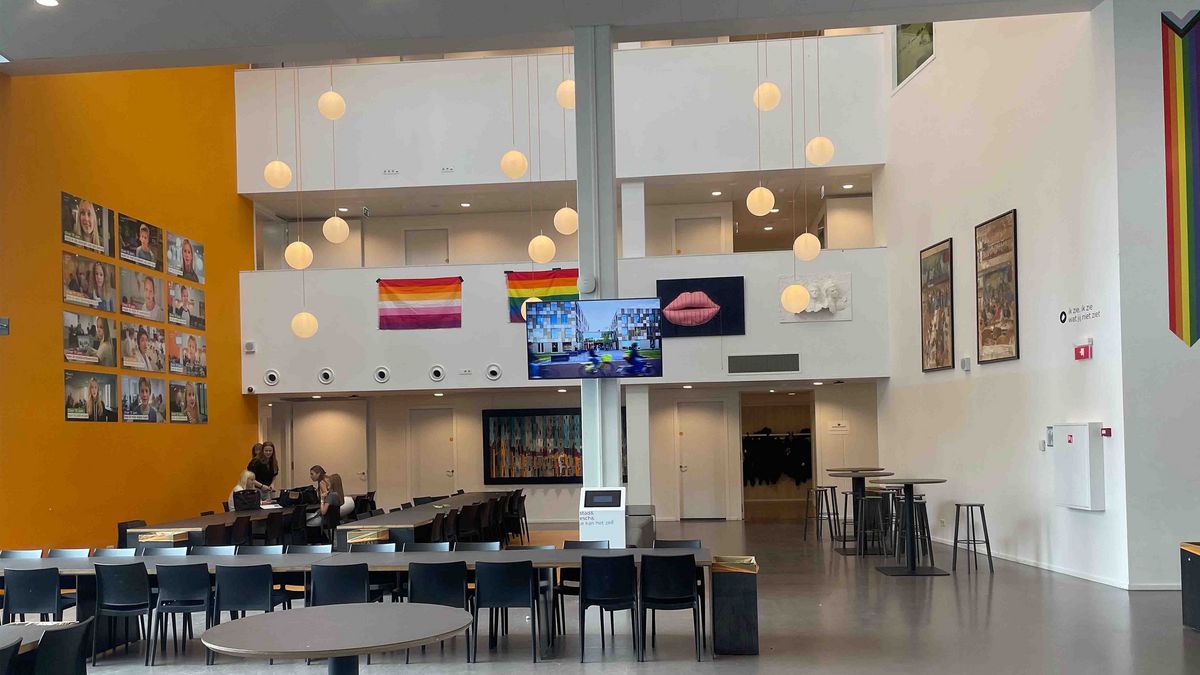 Teaching Across Borders Ein großer, heller Flur in der Schule Stad&Esch mit Porträts, Kunstwerken und Regenbogenflaggen. Im Mittelpunkt ein großer Bildschirm mit Video.