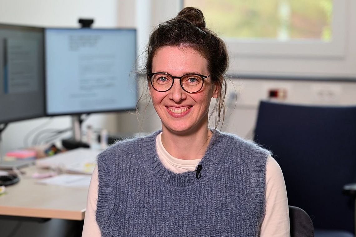 Solveig Elias lächelt in die Kamera. Sie sitzt in ihrem Büro und trägt einen blauen Pullunder, eine Brille und einen Dutt. Im Hintergrund sind Computermonitore zu sehen.