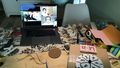 A desk with an open laptop. Next to it are materials such as strings, sponges, glue, scissors, fabric.