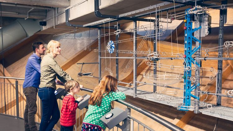 Eine vierköpfoge Familie steht an einem Gitter und probiert eine riesige Kugelbahn aus, die sich dahinter befindet.