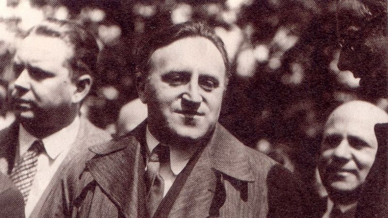 Das Bild zeigt Carl von Ossietzky neben anderen Männern. Er trägt einen Anzug, Krawatte und Mantel.