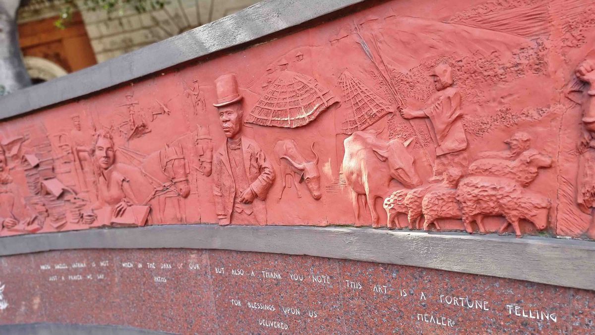 These Walls Ein rotes Relief an einer Wand mit Figuren und Szenen. Unterhalb des Reliefs ein eingravierter Text.