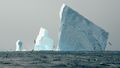 Photo of three icebergs, on the left a small elongated one, in the middle a larger, stepped one and on the right a large, cuboid one. The sky is grey, the sea in the foreground is agitated, the waves carry whitecaps. Birds fly over the water. In front of the larger iceberg, the blow of a fin whale can be seen in the distance. 