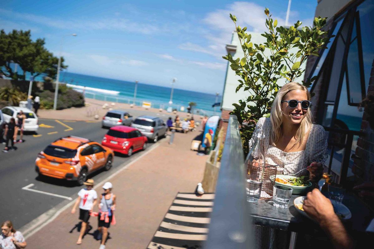 Dunedin Eine Person isst in einem Café und schaut lächelnd eine andere Person an. Im Hintergrund ein belebter Küstenstreifen.