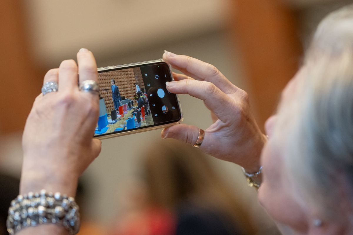 Symposium Das Bild zeigt die Hände einer Person, die ein Smartphone benutzt, um ein Foto zu machen. Auf dem Bildschirm des Smartphones ist ein Bild von einem Innenraum mit Menschen zu sehen. Die Person trägt mehrere Ringe an ihren Fingern und mehrere Armbänder. Das Bild vermittelt den Eindruck, dass jemand einen Moment des Symposiums festhält. Der Fokus liegt auf dem Smartphone und dem Prozess des Fotografierens.