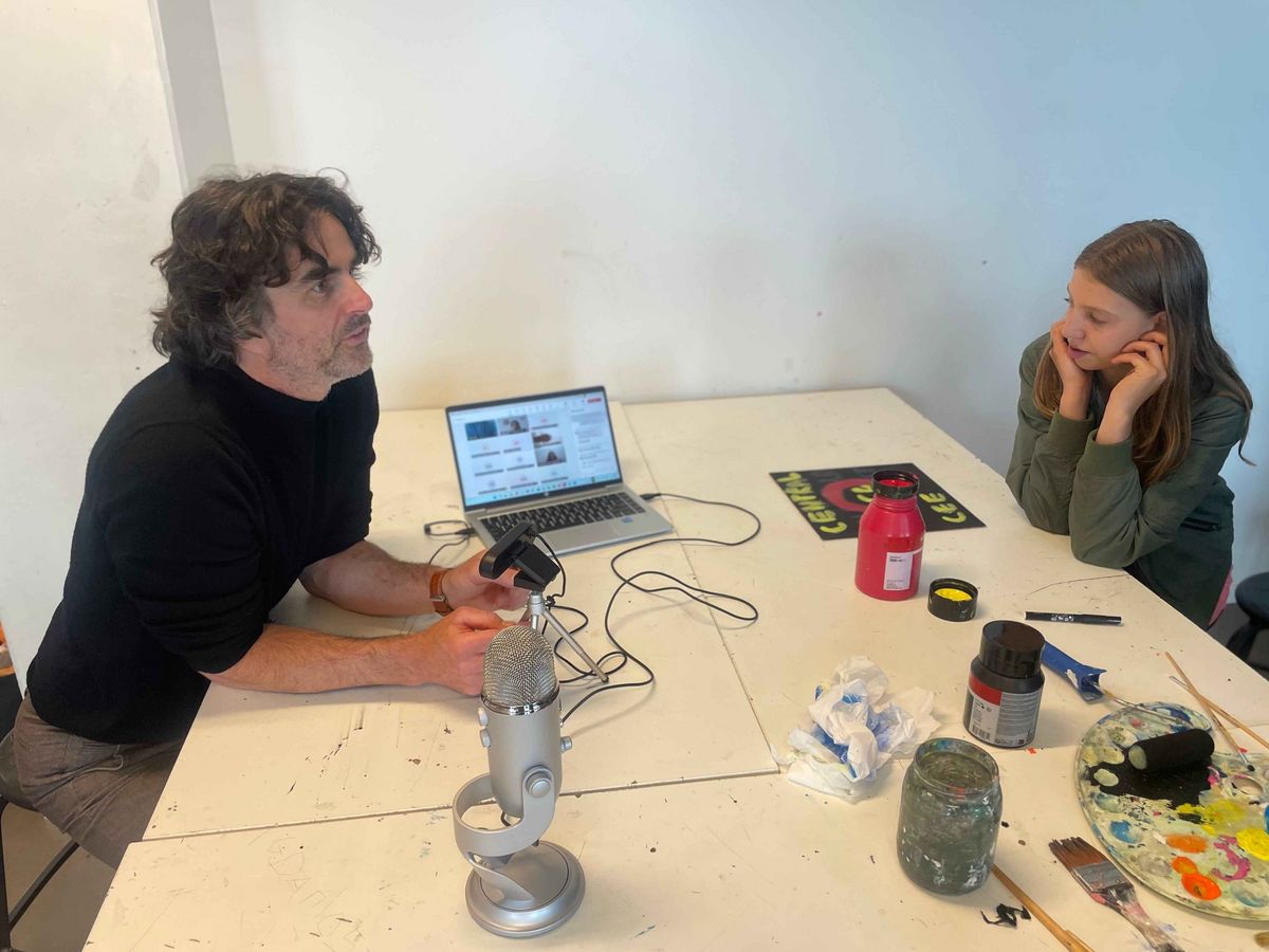 Teaching Across Borders Ein Mann und ein Mädchen sitzen an einem Tisch und arbeiten. Der Mann spricht in ein Mikrofon, das Mädchen ist nachdenklich.