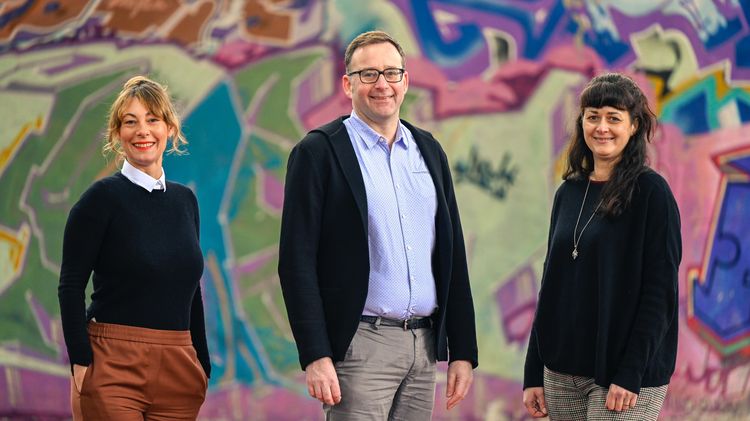 Die drei Personen stehen vor einer Wand mit buntem Graffiti.