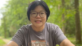 The picture shows Hongyan Bao. She is sitting on a bicycle and smiles into the camera. A forest and a cycle path can be seen in the background.