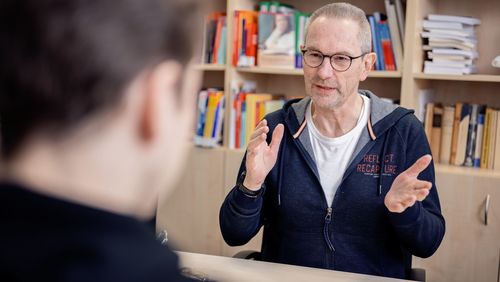 The picture shows Clemens Hillenbrand in conversation with the interviewer. Hillenbrand is sitting in front of a bookshelf and is gesticulating.