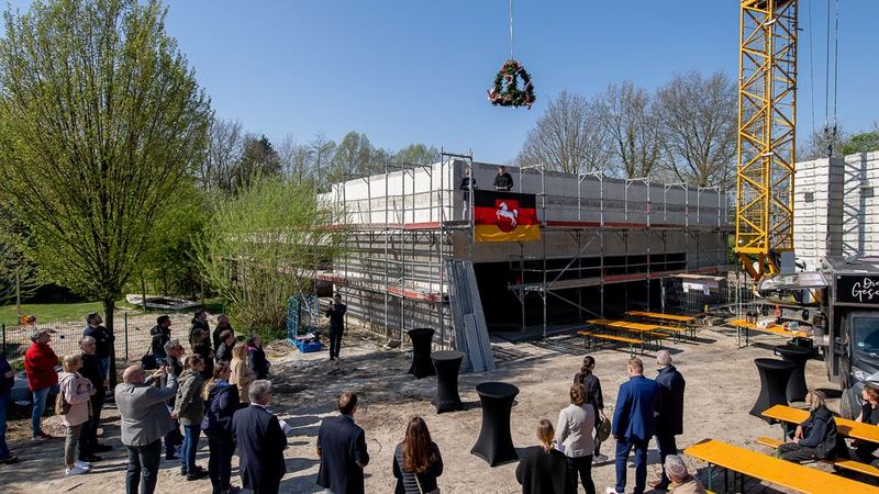 Totale auf den Rohbau, über dem der Richtkranz hängt. Einige Leute stehen um das Gebäude herum.