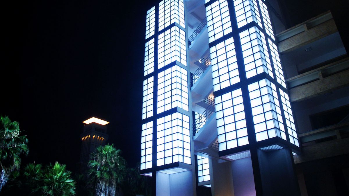 Das Bild zeigt ein modernes Gebäude der National Dong Hwa University (NDHU) bei Nacht, beleuchtet von zahlreichen Lichtern. Das Gebäude besteht aus mehreren rechteckigen Segmenten, die durch große Fenster und eine geometrische Anordnung von Lichtquellen hervorgehoben werden. Die Beleuchtung erzeugt einen auffälligen Kontrast zum dunklen Nachthimmel und betont die architektonischen Details des Gebäudes. Im Vordergrund sind einige Palmen zu sehen, die dem Bild eine tropische Atmosphäre verleihen.