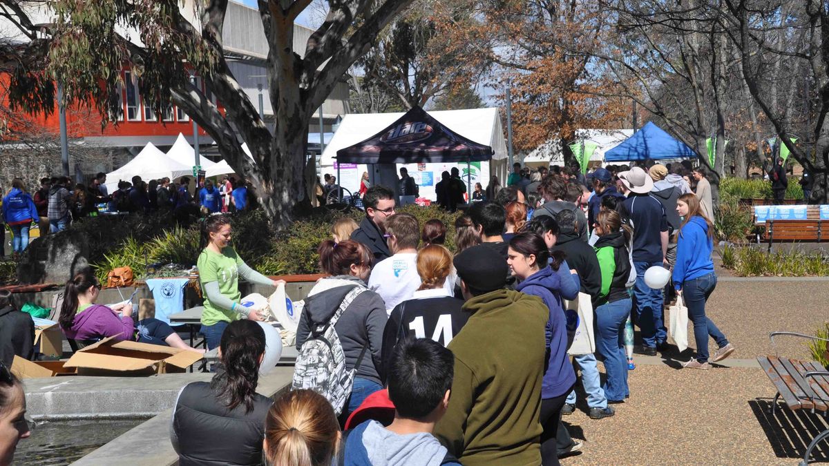 Das Bild zeigt eine belebte Veranstaltung auf dem Campus der University of New England. Viele Menschen sind versammelt, einige stehen in Schlangen vor Ständen, andere sitzen. Im Hintergrund sind Zelte und Gebäude der Universität zu sehen. Die Atmosphäre wirkt festlich und gesellig.