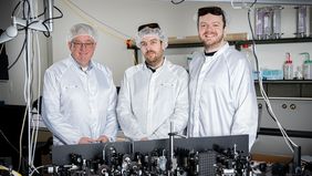 Drei Männer stehen in einem Labor. Alle drei tragen weiße Kittel, eine Haube über den Haaren und eine spezielle Brille, die die Augen vor dem Laserlicht schützt. Im Vordergrund ist der Versuchsaufbau auf einem Tisch zu sehen.