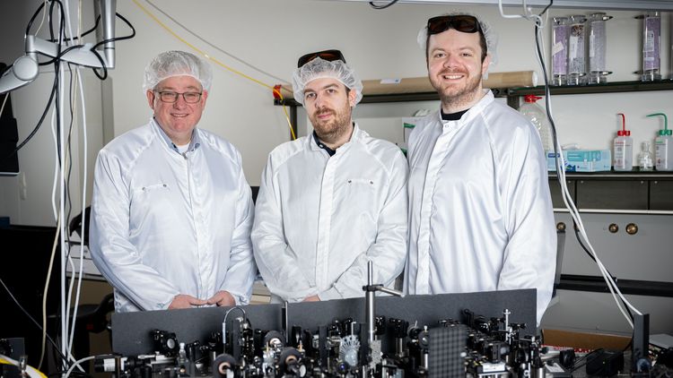 Drei Männer stehen in einem Labor. Alle drei tragen weiße Kittel, eine Haube über den Haaren und eine spezielle Brille, die die Augen vor dem Laserlicht schützt. Im Vordergrund ist der Versuchsaufbau auf einem Tisch zu sehen.