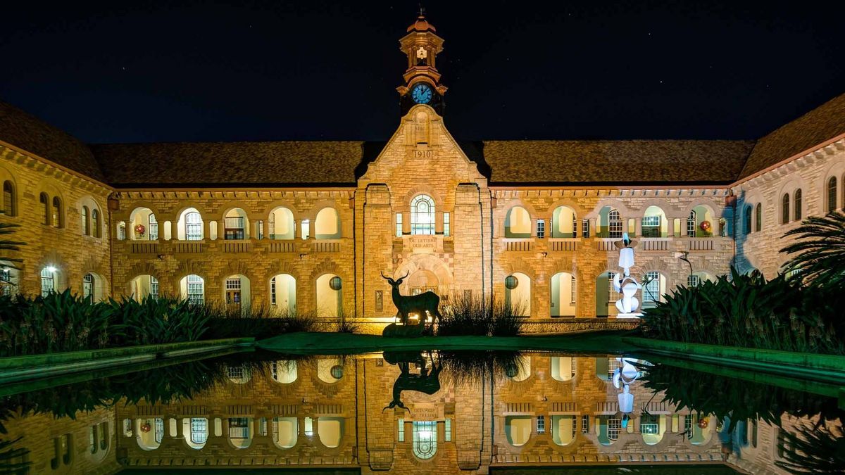 Ein historisches Gebäude des Hatfield Campus der University of Pretoria ist nachts beleuchtet und spiegelt sich in einem Teich vor dem Gebäude wider. Das Gebäude ist aus hellem Stein gebaut und verfügt über einen Uhrenturm. Die Beleuchtung betont die architektonischen Details und erzeugt eine ruhige Atmosphäre. Vor dem Gebäude steht eine Statue, augenscheinlich eines Springbocks, einer südafrikanischen Antilopenart. 