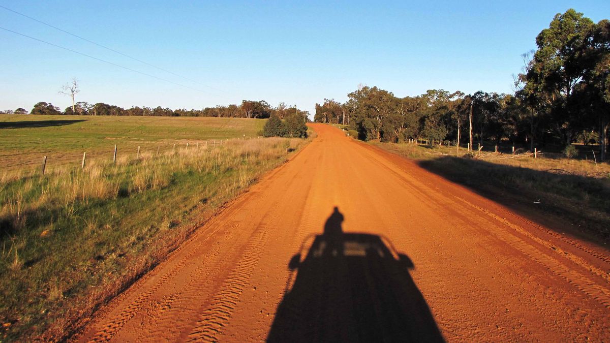 Das Bild zeigt eine lange, gerade Landstraße, die sich durch eine leicht hügelige Landschaft erstreckt. Die Straße ist rotbraun und wird von grünen Feldern und Bäumen gesäumt. Im Vordergrund ist der Schatten einer Person zu sehen, die in einem Fahrzeug sitzt und in die Ferne blickt. Der Himmel ist blau und die Sonne scheint. Die Szene vermittelt ein Gefühl von Freiheit und Abenteuer.