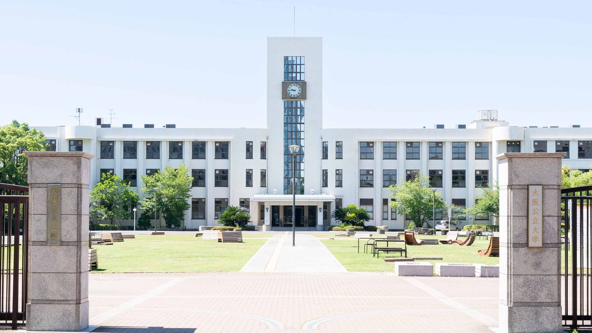 Das Bild zeigt einen zentralen, weißen Gebäudekomplex der Osaka Metropolitan University mit einem markanten Uhrturm. Der Bau ist rechteckig und verfügt über mehrere Stockwerke mit zahlreichen Fenstern, die eine symmetrische Fassade bilden. Vor dem Gebäude erstreckt sich ein gepflegter Rasen mit Bäumen und Bänken, der durch einen Weg zum Haupteingang führt, der von zwei steinernen Säulen mit japanischen Schriftzeichen flankiert wird.