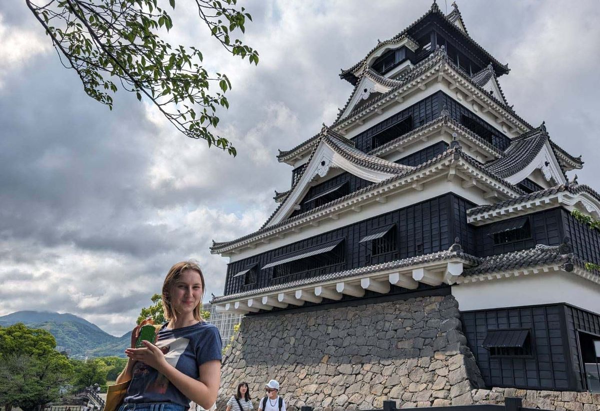 Burg Kumamoto ist eine mehrstöckige Burg mit dunklen Holzaußenwänden und weißen Putzdetails. Eine Person steht im Vordergrund und posiert lächelnd mit einem Getränk in der Hand. Steinmauern und Grünpflanzen sind um die Burg herum sichtbar.