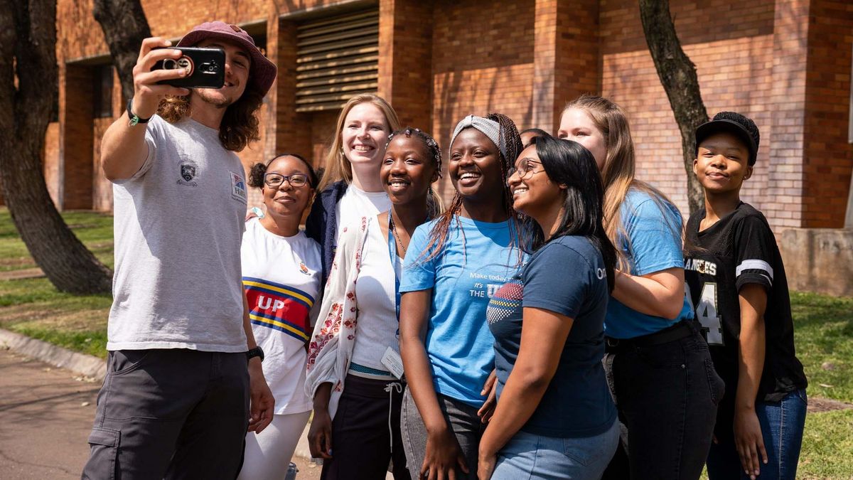 Eine Gruppe von neun jungen Menschen steht auf dem Campus der University of Pretoria zusammen und macht ein Selfie mit einem Smartphone. Sie lächeln und wirken fröhlich. Einige tragen Kleidung mit dem Logo der Universität. Im Hintergrund befindet sich ein Backsteingebäude und Bäume.