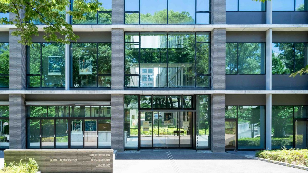 Das Bild zeigt die Fassade eines modernen, mehrstöckigen Gebäudes der Osaka Metropolitan University, umgeben von üppigem Grün. Die Fassade besteht aus grauen Betonplatten und großen Glasfenstern, die das Licht reflektieren und einen Einblick in die Innenräume gewähren. Vor dem Eingangsbereich befindet sich eine niedrige Mauer mit sowohl japanischen als auch englischen Schriftzügen, die verschiedene Institute ausweisen.