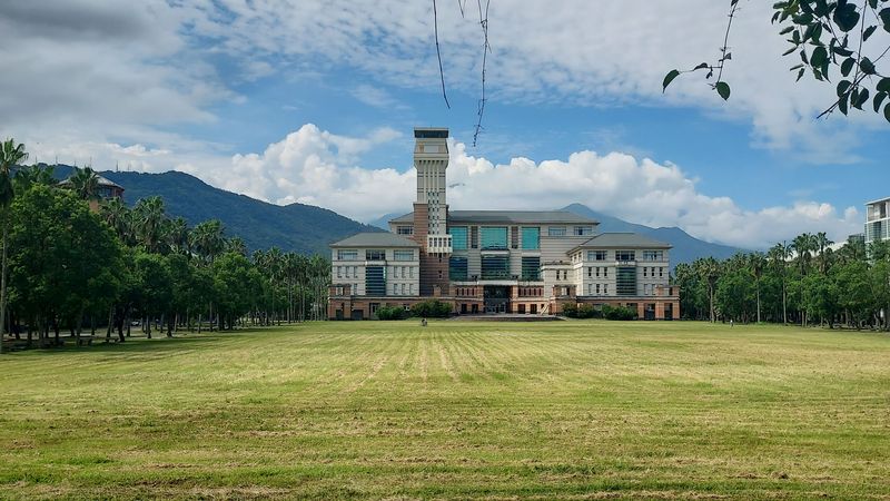 Das Hauptgebäude der Universität hinter eine gemähten WIese, an der Seite sind Bäume zu sehen und im Hintergrund bewaldete Berge.. 