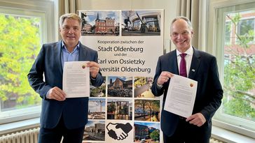 Zwei Männer stehen vor einem Aufsteller und halten ein Blatt Papier in die Kamera. 
