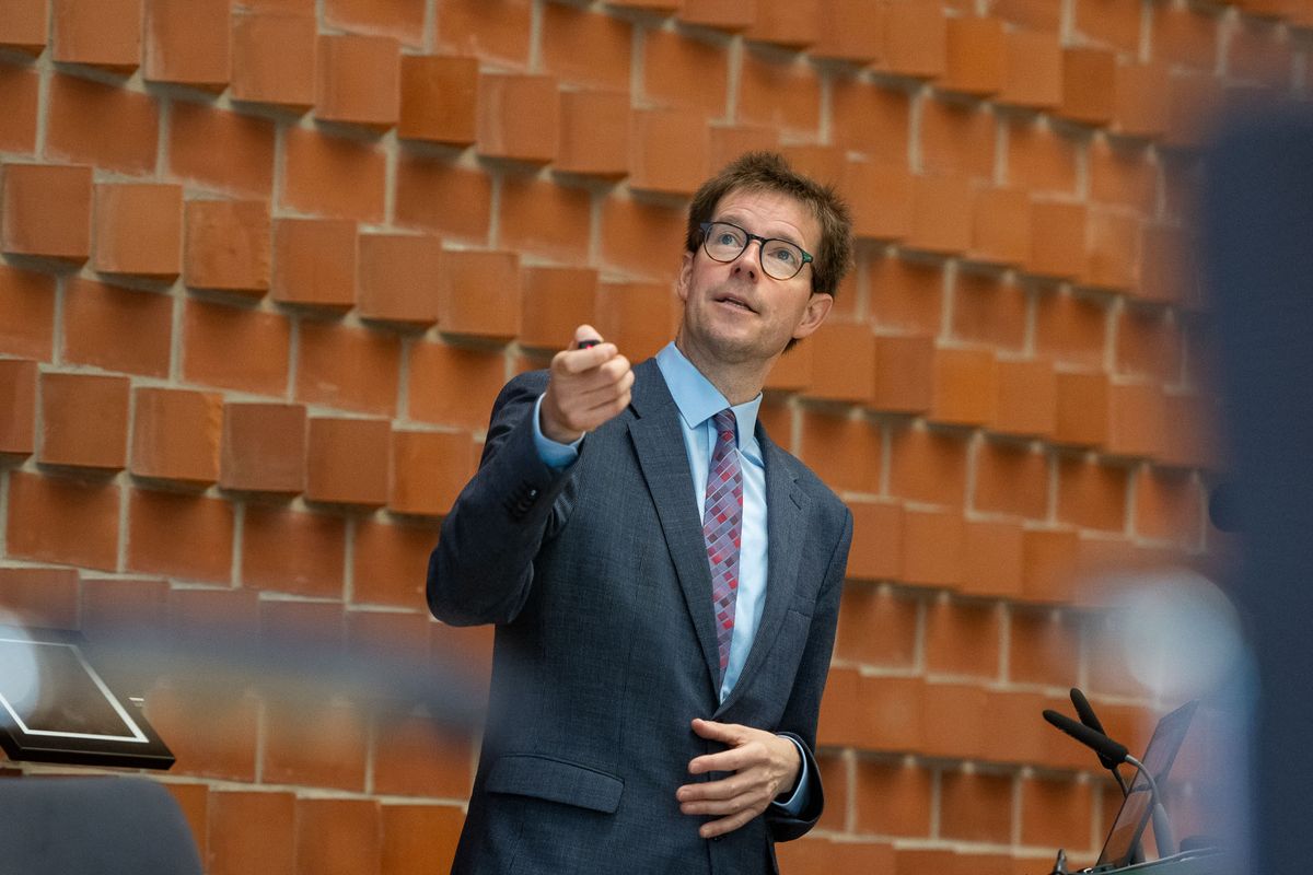 Symposium Ein Mann in einem grauen Anzug mit bunter Krawatte steht vor einer roten Backsteinwand und hält eine Rede. Er trägt eine Brille und deutet mit der rechten Hand auf etwas außerhalb des Bildes. Ein Mikrofon steht vor ihm auf einem Pult.