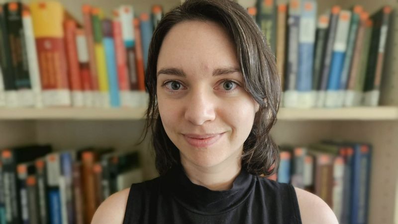 Das Bild zeigt Christina-Marie Juen. Sie sitzt vor einer Bücherwand und schaut in die Kamera.