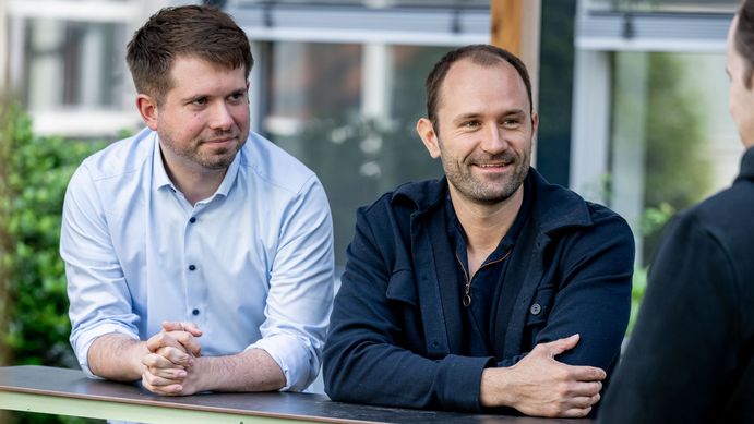 Das Bild zeigt Tim Wohlann und Leon Hoffmann, die sich mit dem Interviewer unterhalten. Im Hintergrund ist das Gebäude V03 zu erkennen. Wohlann lächelt den Interviewer an.