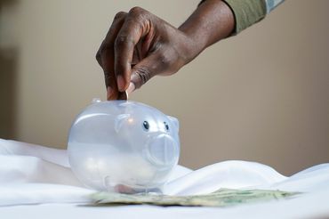 Auf dem Bild ist ein durchsichtiges Sparschwein zu sehen, in das eine Hand eine Münze steckt.