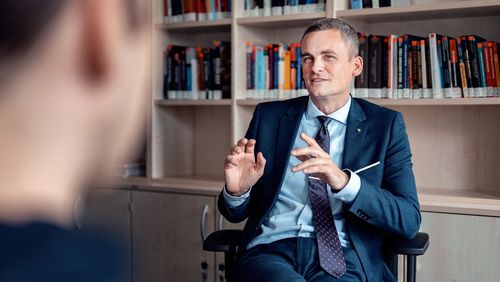 Das Bild zeigt Philipp Köker. Er sitzt an seinem Schreibtisch vor einem Bücherregal und spricht mit dem Interviewer. Mit seiner linken Hand gestikuliert er leicht.