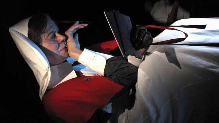 The picture shows a waxwork nurse lying under a blanket in a tent or house. She is reading something. She also appears to be crying and appears to be wiping a tear from her face with her right hand. The scene is very dark.