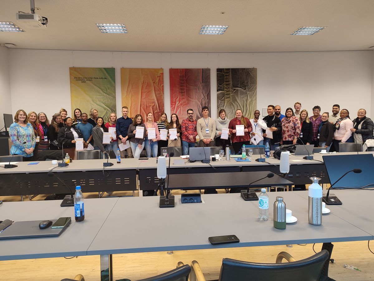 Teilnehmende der DAAD Winter School posieren zusammen mit den Dozierenden und Organisator*innen der DAAD Winter School. Die Gruppe lächelt in die Kamera, die Teilnehmenden präsentieren ihre Abschlusszertifikate. Im Vordergrund sind Tische und Stühle mit Wasserflaschen und Tagungsequipment zu sehen.  