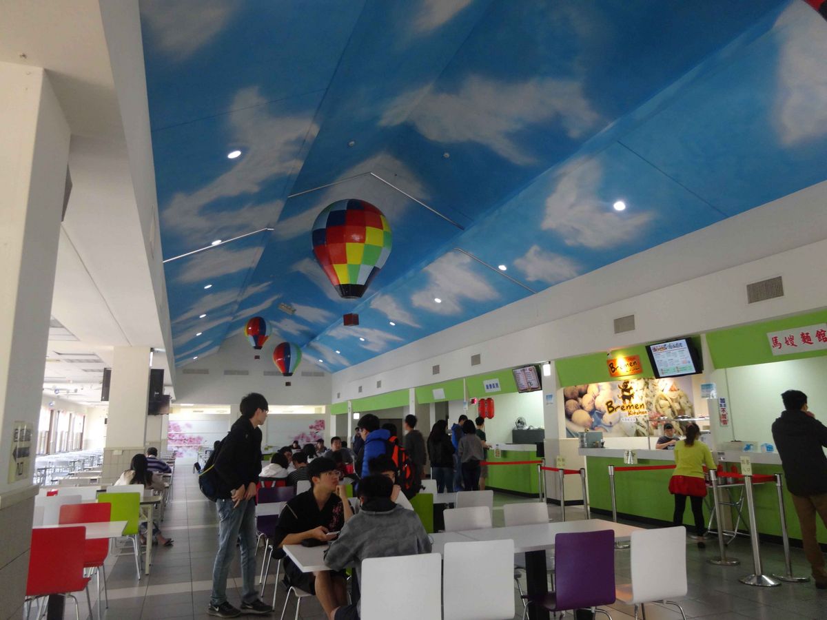 Das Bild zeigt eine belebte Mensa mit einer fantasievollen Deckenbemalung, die einen blauen Himmel mit herunter hängenden Heißluftballons darstellt und eine fröhliche Atmosphäre schafft.