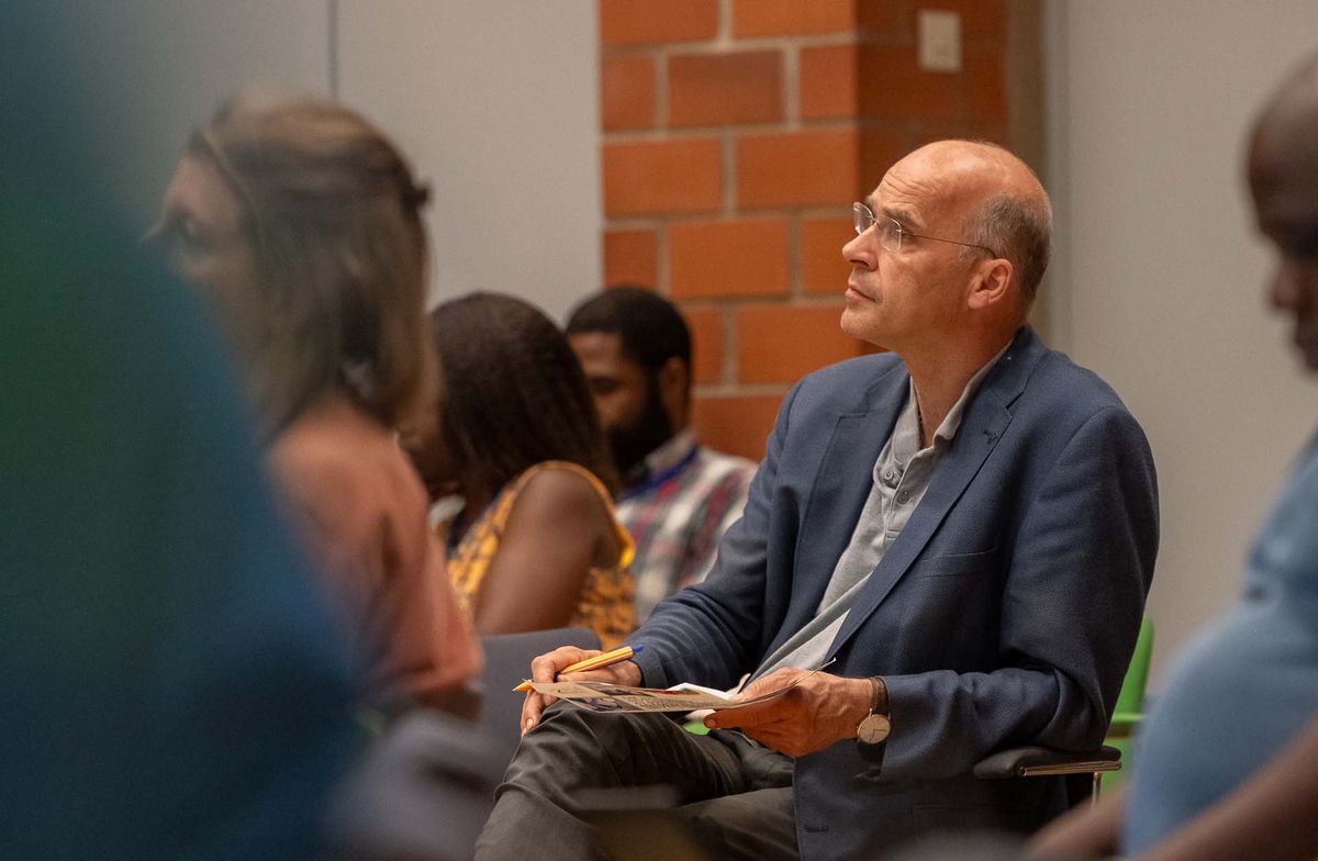 Symposium Ein Mann mit Brille sitzt auf einem Stuhl und blickt nach vorne. Er trägt einen blauen Anzug und hält ein Blatt Papier und einen Stift in seinen Händen. Im Hintergrund sind verschwommene Personen und eine rote Backsteinwand zu sehen. Das Bild vermittelt den Eindruck, dass der Mann aufmerksam einer Präsentation oder Diskussion zuhört. Er scheint Notizen zu machen oder sich auf das Gehörte zu konzentrieren.