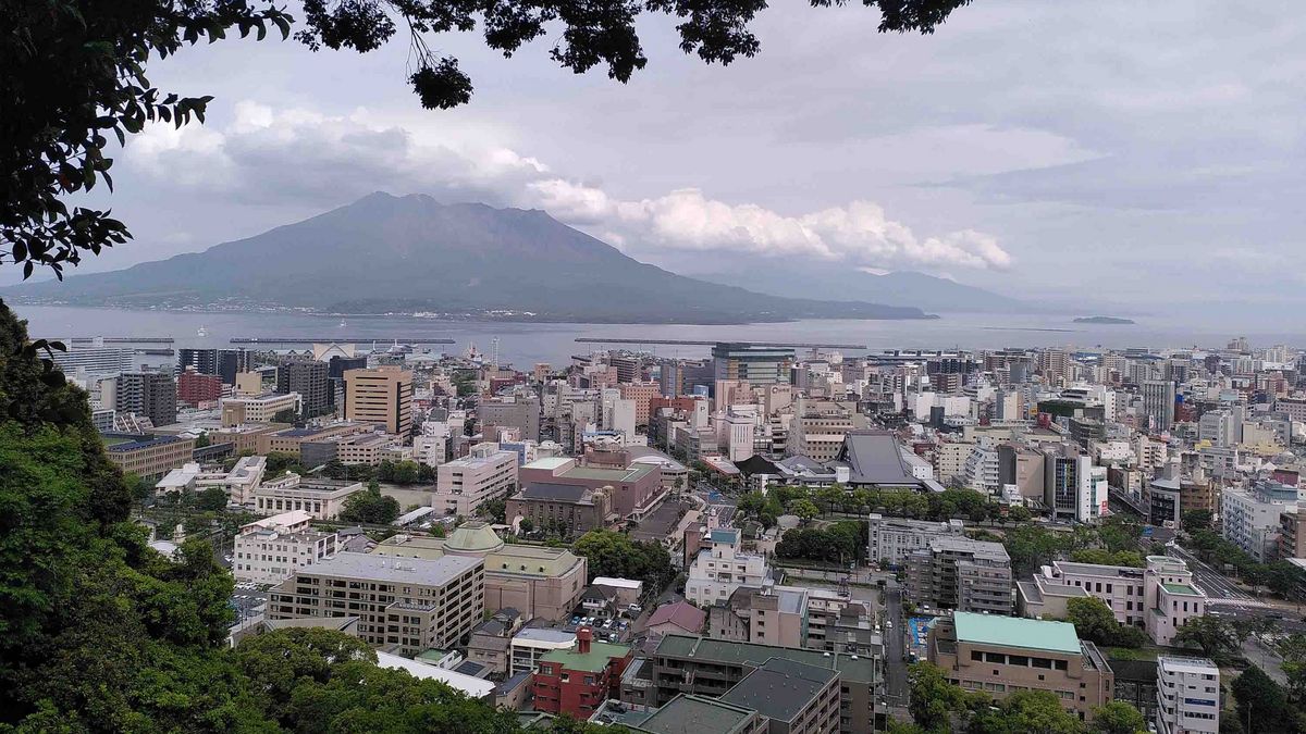 Ein Blick über Kagoshima Stadt mit dem Sakurajima Vulkan im Hintergrund. Im Vordergrund sind Bäume und Gebäude zu sehen, dahinter erstreckt sich die Stadt bis zur Kagoshima Bucht. Der Himmel ist bewölkt.