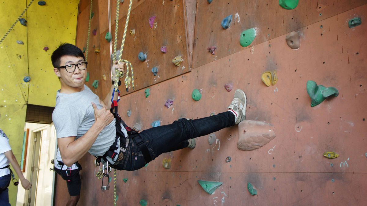 Das Bild zeigt einen Studierenden, der an einer Kletterwand klettert. Der Studierende trägt eine Kletterausrüstung, darunter ein Seil, einen Gurt und Kletterschuhe und befindet sich in einer dynamischen Kletterposition. Im Hintergrund ist ein Teil der Kletterwand mit bunten Griffen zu sehen. Das Bild vermittelt den Eindruck, dass die NDHU ihren Studierenden Möglichkeiten für sportliche Aktivitäten und Freizeitgestaltung bietet.