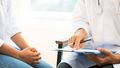 A young woman sits next to a doctor who shows something on a clipboard. The hands are in the center of the picture.