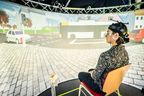 Eine Frau mit Hörgerät sitzt vor einer Leinwand, auf der ein Straßenverkehr dargestellt wird.