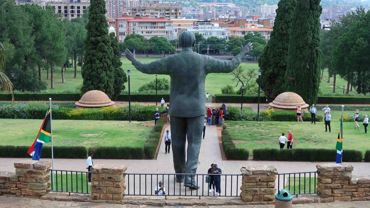 Das Bild zeigt eine große Statue von Nelson Mandela, dargestellt mit ausgebreiteten Armen. Die Statue steht in einem Park mit gepflegten Grünflächen, Bäumen und Wegen. Im Hintergrund sind Gebäude und eine städtische Landschaft zu sehen. Mehrere Personen sind im Park zu sehen, die die Statue betrachten oder passieren. Im Vordergrund ist die südafrikanische Flagge zu erkennen.
