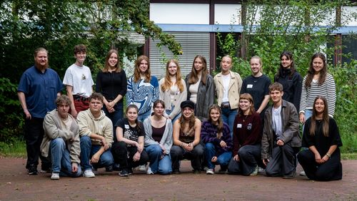 Das Bild zeigt 19 der 22 Freiwilligen, die sich zu einem Gruppenbild aufgestellt haben. Sie stehen vor einem Unigebäude und lächeln in die Kamera.